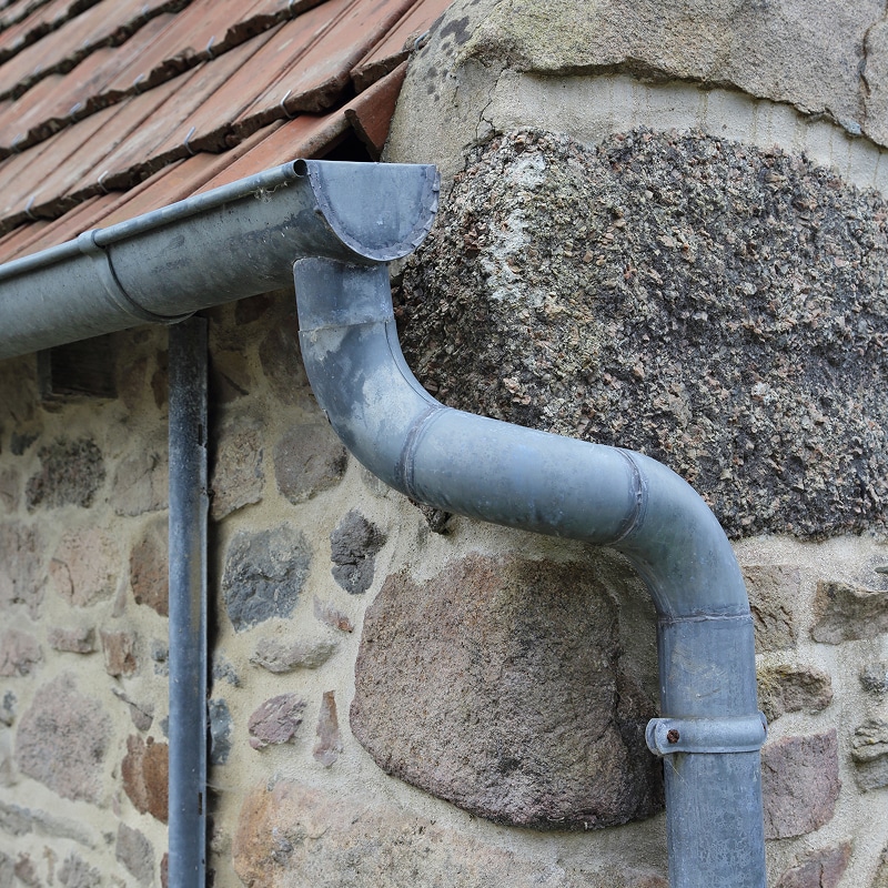 Gouttière et descente pluviale en zinc vieilli contre un mur de pierres apparentes avec une corniche en tuiles rouges.