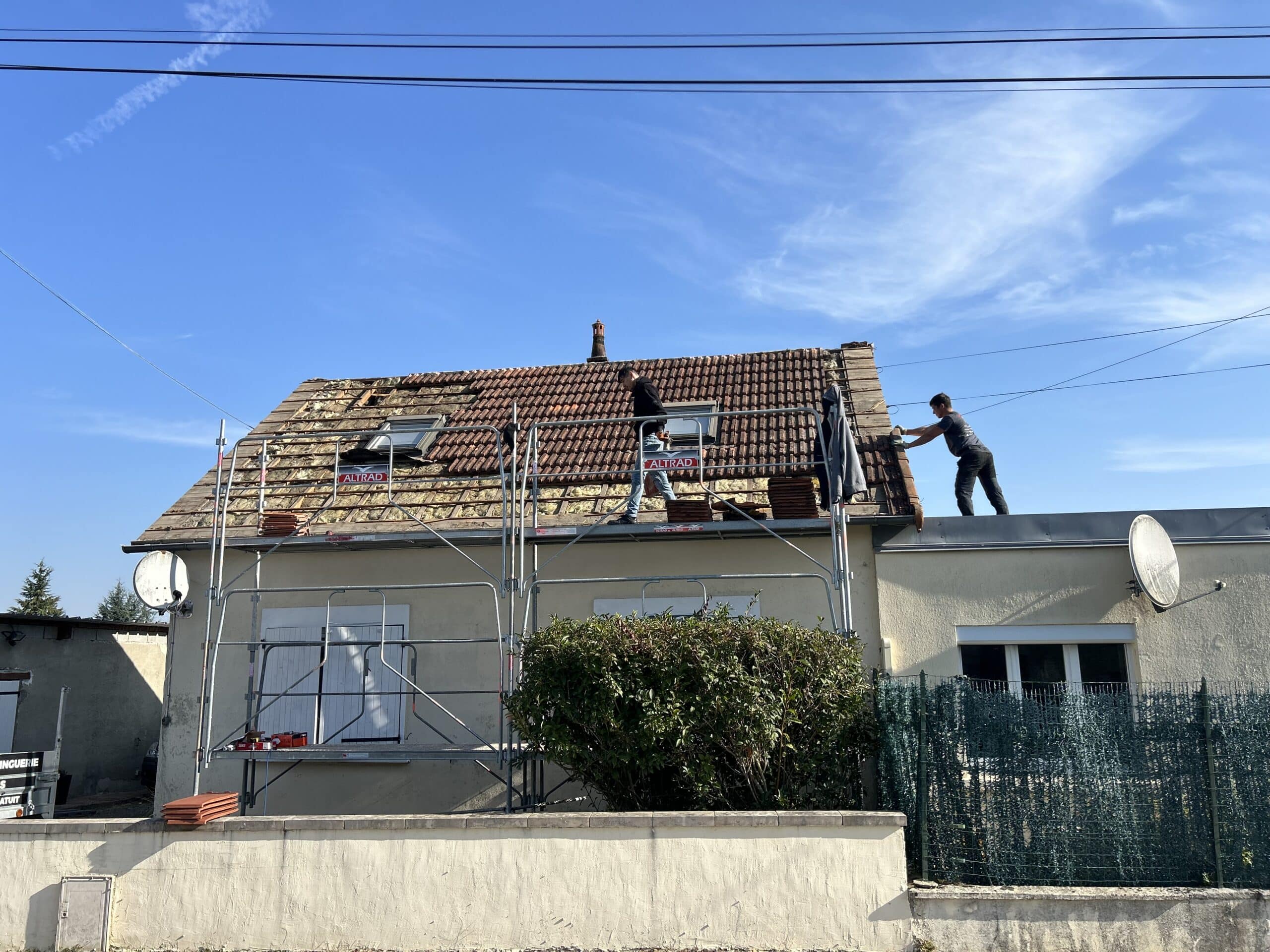 Trois ouvriers effectuent des travaux de rénovation de toiture avec tuiles, utilisant un échafaudage Altrad sous un ciel bleu dégagé.