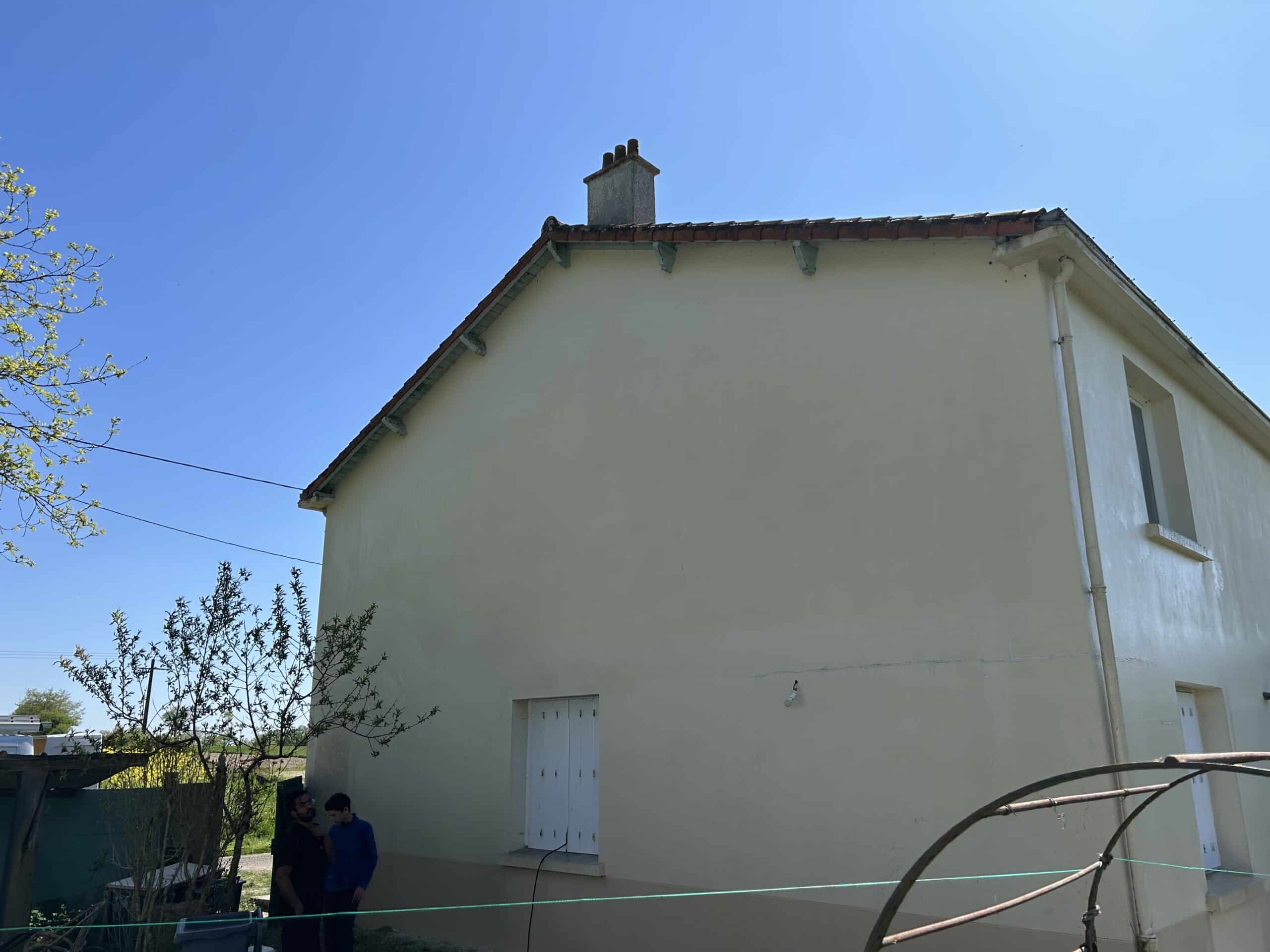 Façade latérale d'une maison crépie beige sous un ciel bleu vif, avec une souche de cheminée et des poutres de toit décoratives. Deux personnes sont visibles à gauche.