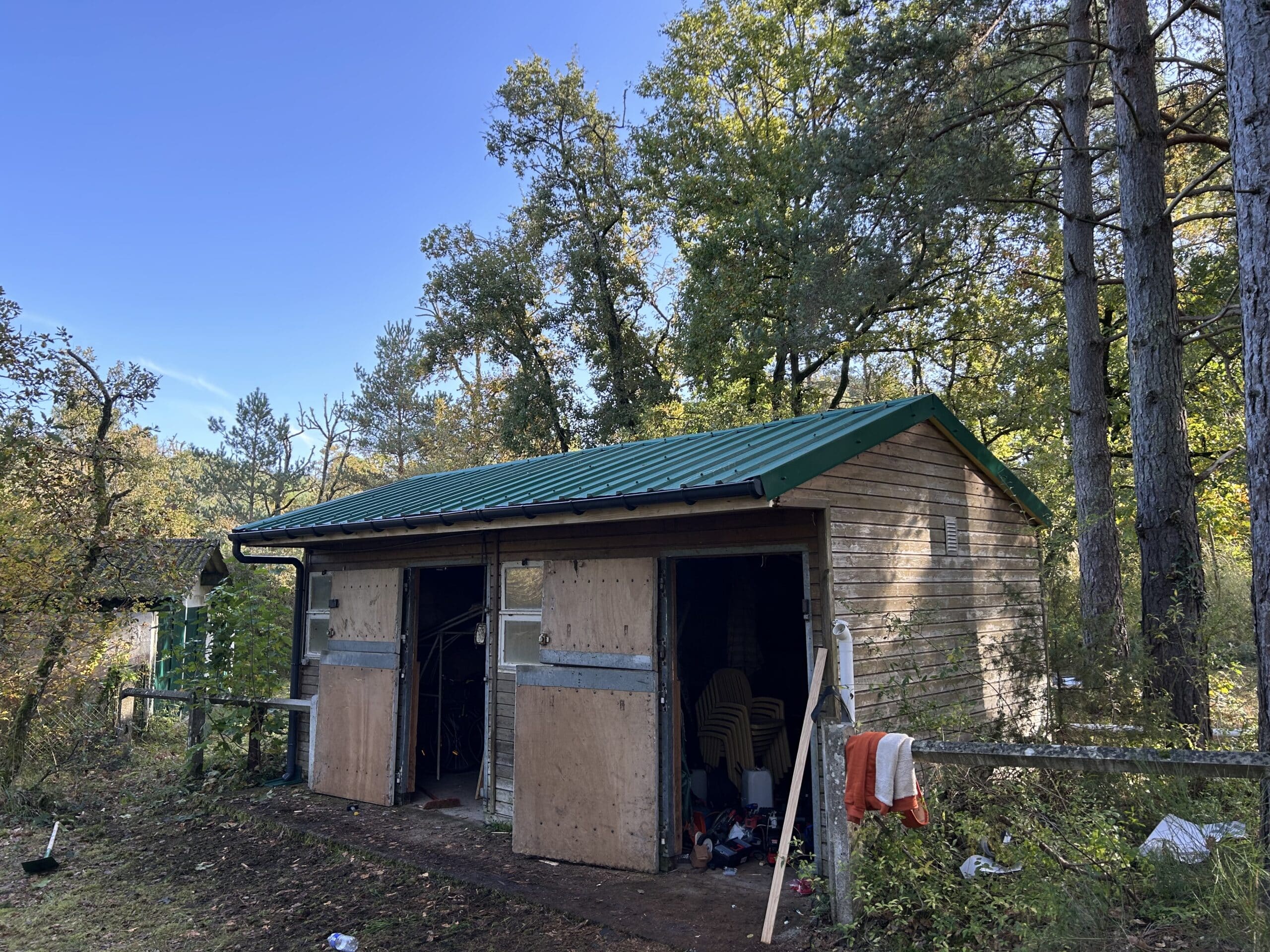 Cabane en bois avec toit vert ondulé, portes ouvertes révélant des chaises empilées, entourée d'arbres feuillus en automne, ciel bleu clair.