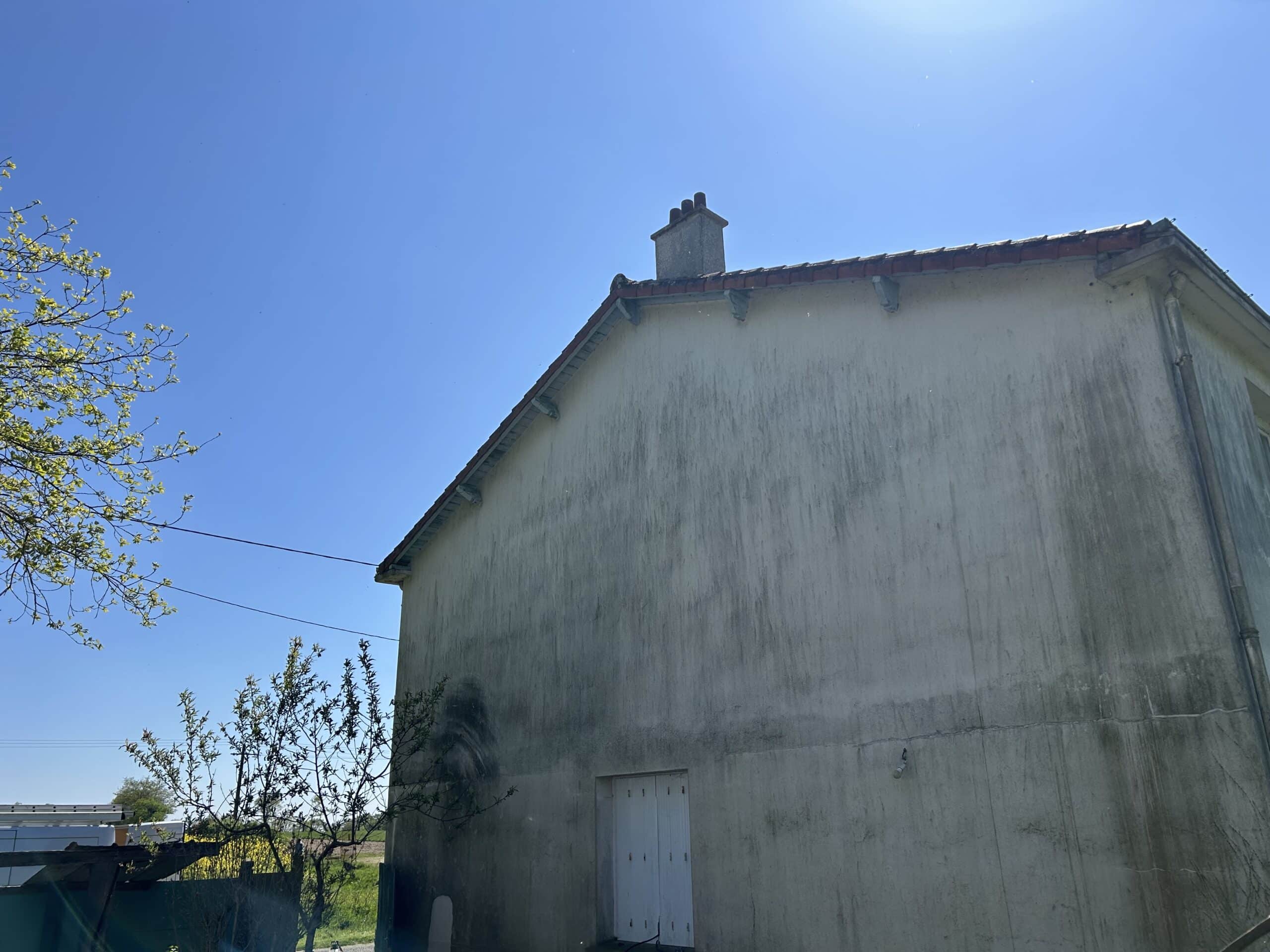 Façade d'une maison ancienne, mur sale avec coulures sombres, sous un ciel bleu clair et ensoleillé, branches d'arbre à gauche.