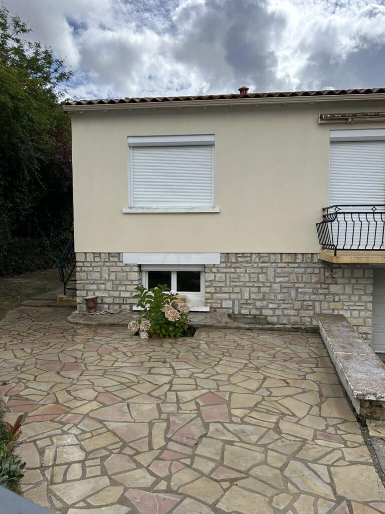 Façade de maison claire avec soubassement en pierres, volets roulants blancs fermés et terrasse en dalles irrégulières aux tons ocres et rosés.