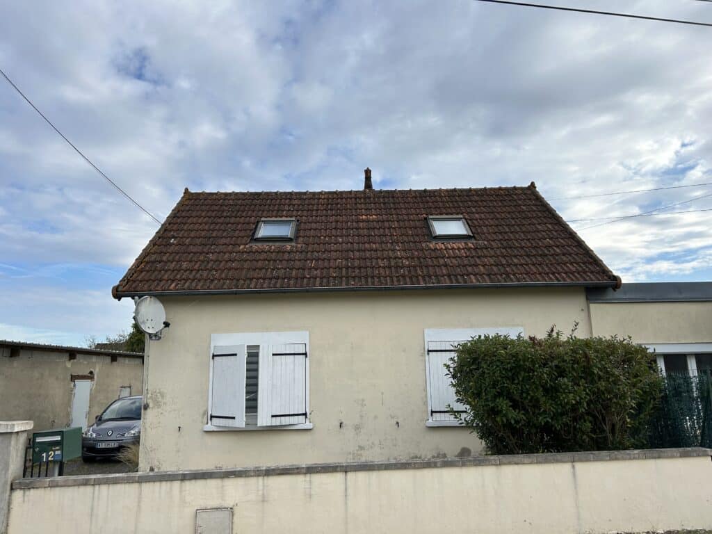 Façade d'une maison de plain-pied avec murs clairs, toit en tuiles rouges, deux lucarnes et volets blancs, plaque '12 BIS'.