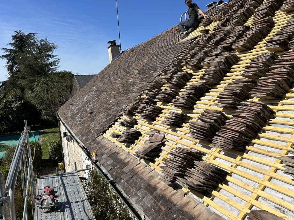 Rénovation de toit en tuiles anciennes : couvreur travaillant sur liteaux jaunes, tuiles empilées. Scaffolding et scie Milwaukee visibles.